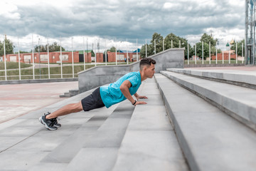 male athlete is pushing up from concrete steps in summer city. Fitness motivation youth lifestyle. Free space for copy text. Background stairs, clouds. Sportswear. T-shirt. Shorts and Sneakers.
