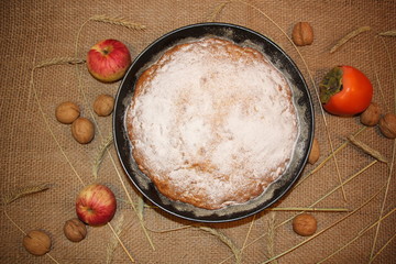 Homemade pie with apple, persimmon, walnuts, sugar powder, grains in baking pan on sackloth rustic ...