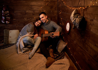 Fototapeta na wymiar Young couple in love on romantic date in wooden room hut on christmas new years eve. Brunette man, wearing grey winter sweater, holding playing guitar for pretty woman covered with warm blanket.