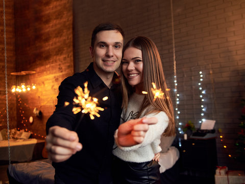 Young couple in love, holding sparklers, smiling laughing celebrating new year in dark room. Handsome brunette man, wearing black shirt with pretty woman in white sweater on New years Christmas eve.