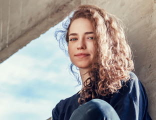 Lovely girl in dark clothes sits on concrete block in industrial zone. Face close-up