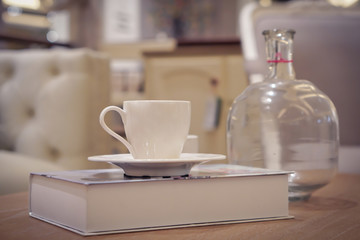 Focus selection: A white coffee cup placed on a book at a table and a glass bottle near it. In a light, comfortable atmosphere