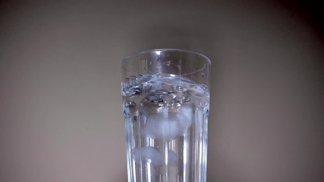 Slow motion close shot of a hand dropping ice cubes into a tall glass of water, then pouring concentrated orange juice from a plastic bottle. 