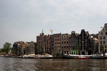 View of Amstel river, canal boats and historical, traditional and typical buildings reflecting Dutch architectural style in Amsterdam. It is a summer day with cloudy sky.