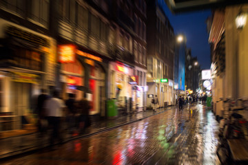 Fototapeta na wymiar Blurry motion image of people walking on Warmoesstraat street in Amsterdam. It is one of the main shopping streets with cafes, restaurants and shops. It is a rainy summer night. Youth culture concept