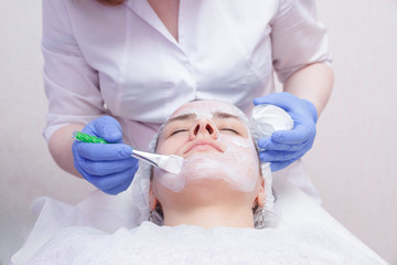 Crop master applying white mask with brush on face and neck of young woman in towel lying on table