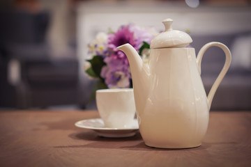 Focus selection: a white coffee pot placed on the table with a coffee cup and flowers as a blurred background