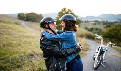 A cheerful senior couple travellers with motorbike in countryside, hugging.