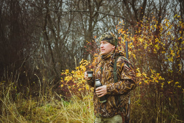 A man in camouflage clothing with a thermos and a gun in a case.