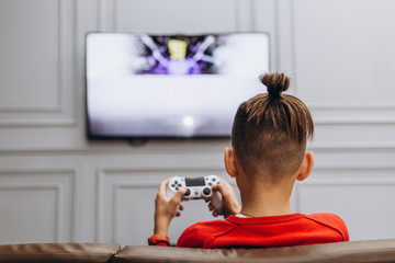 rear view: little school boy sitting in front of big monitor and playing game console one, concept of gadget addiction