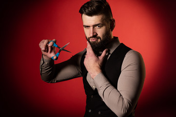 Bearded man in vest on red background. Men cut their beard with hairdressing scissors