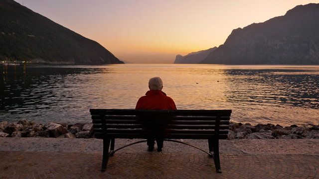 Lonely old man sits on a wooden bench at dusk looking at the lake and the light on the horizon.