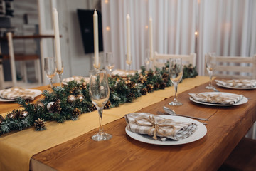 Beautiful Christmas serving table with candles and fir-tree decoration at cozy modern interior close-up. Celebrating december holiday new year surrounded by seasonal decorated glasses and plates
