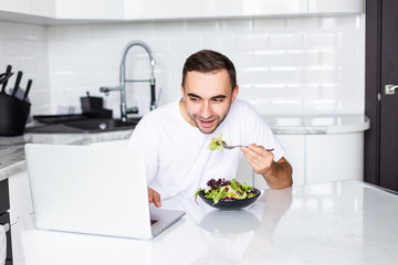 Portrait of handsome man have breakfast while use laptop in white kitchen.