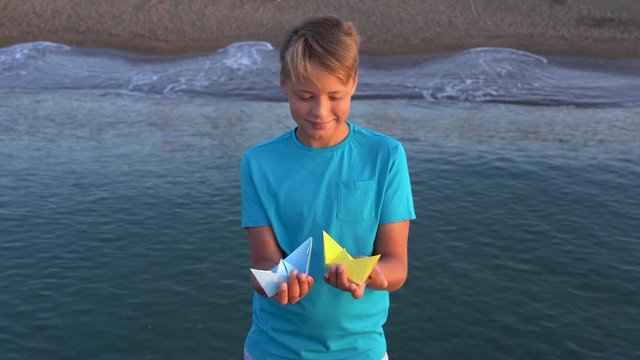 Closeup view of cute handsome white kid holding two paper bright blue and yellow colors ships standing at sunny morning beach. Happy summer tourism concept. Real time 4k video footage.