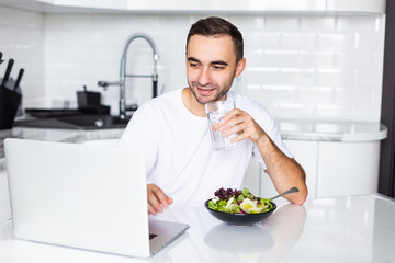 Fototapeta na wymiar Young man wearing casual clothing eating breakfast and drinking water at home while using laptop