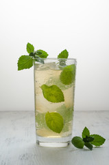 The glass of ice cold transparent beverage in which float the mint leaves. A mint leaf sits next to a glass on a white wooden table.