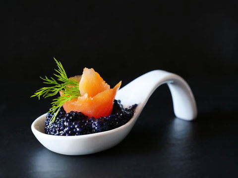 Black caviar with smoked salmon bite in white porcelain spoon over black background.