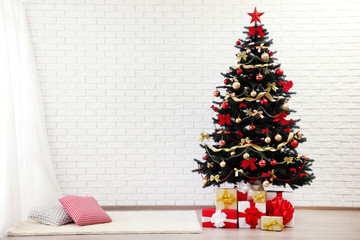 Christmas fir tree with ornaments and gift boxes standing at home near white brick wall