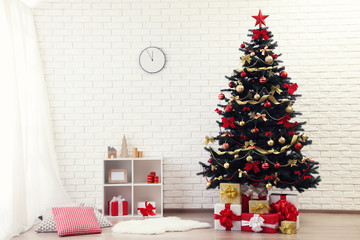 Christmas fir tree with ornaments and gift boxes standing at home near white brick wall