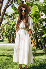 Obraz na płótnie Canvas stylish young attractive woman dressed in a long linen dress with a hat photographed in the summer on a background of palm trees and the sea in the bright sun
