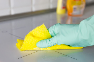 Close up of woman hand in protective rubber gloves washing or cleaning modern white induction hob by a rag in the kitchen, blurred cleaning products on background. 