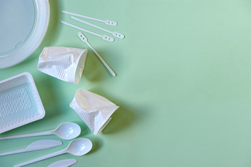 Two smashed white plastic coffee cups, spoons, knives, stirrers, plate and box on a light green background copy space and soft shadows. Zero waste, plastic free, stop pollution, ecological concept.