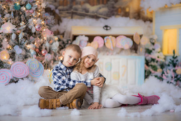 fair-haired happy cute children brother and sister 6-7-8 years near the Christmas tree are sitting in an embrace, on the eve of the  and Christmas holiday bright colors, pink and blue, square street