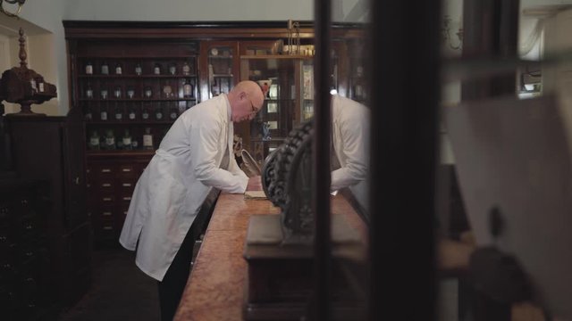 Side view of old energetic Caucasian man in white robe writing with feather pen. Pharmacist staying at cash counter revising his notes. Retro drugstore, vintage pharmacy, pharmaceutical museum.