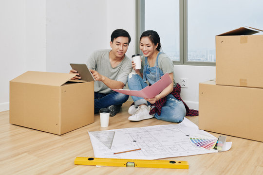 Cheerful young Vietnamese couple discussing construction plans in documents and on tablet computer when planning apartment renovation