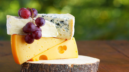 Camembert, brie, hard cheese and blue cheese on wooden board. Slices of various cheeses and grapes on blurred background. Dorblu and soft cheese on wooden boards. Widescreen