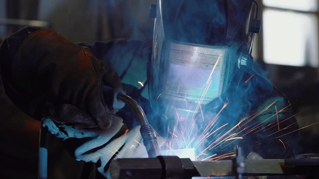 Close up: worker in face shield or protective mask uses gas welding torch to connect the seam of metal part in industrial factory. Blue fire flickers, orange sparks are flying, blacksmith manual work.