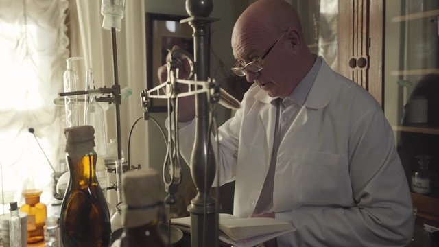 Portrait of old bald Caucasian man checking ancient flasks, containers and test tubes, and writing with feather pen. 19th century pharmacist revising vintage drugstore. Retro style, healthcare