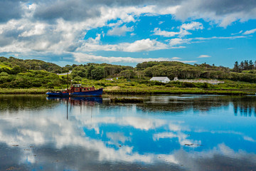 Clouds reflecting in the water, a ship anchored in the water at high tide in Clifden Bay, surrounded by green vegetation, spring day with a blue sky and white clouds in Clifden, Ireland