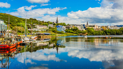 Fototapeta na wymiar Pier at the port of Clifden at high tide, boats anchored with mirror reflection in the water, sunny spring day with a blue sky and abundant white clouds in Clifden, Ireland