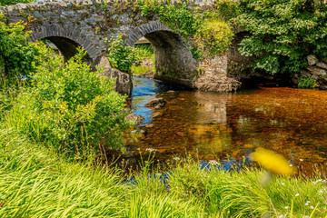 Fototapeta na wymiar Ardbear Old Bridge over the Owenglin or Owenglen river with clean and transparent water surrounded by green vegetation, sunny and calm day in Clifden, Ireland