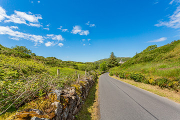 Rural road with limestone fences between hills with green vegetation, sunny spring day with a blue sky and white clouds in the province of Connacht, Ireland