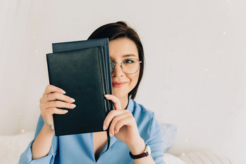 Young woman in glasses with black short hair covered her face with a book