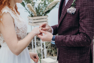 the bride and groom put on each other rings, a burgundy suit and a beige wedding dress, cloudy day, a ceremony by the sea, hands close-up