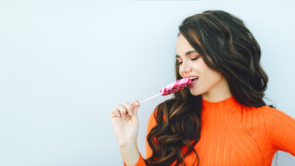 Obraz premium Young beautiful girl with dark curly hair eats a pink lollipop candy screaming proud in a bright orange sweater on a light plain background