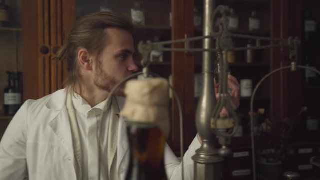 Handsome Caucasian man in white robe looking at old-fashioned bottle with drug, talking and handing flask to young redhead woman. Camera following test tube. Vintage pharmacy, retro, 19th century.