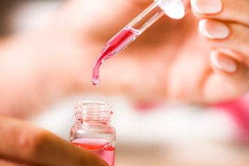 Woman holding transparent glass pipette with pink falling liquid and drop detail in her finger...