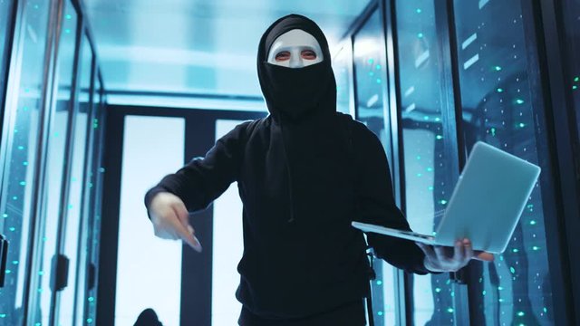 Masked anonymous hacker holding laptop gesturing funny at camera in database. Professional spy breaking server in seconds, cheering up with fist pump inside server room.