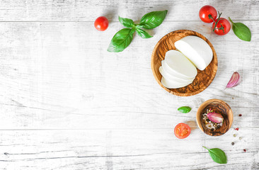 Mozzarella cheese in wooden bowl on white rustic table top view. Soft white italian cheese, tomatoe and basil on old board with copy space..