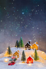 Miniature wooden houses village on the snow at night over blurred Christmas starry decoration background, toned