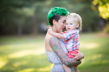 A portrait of Caucasian young woman with green hair and tattoo holding a blond toddler boy in her hands. Hugging, Mother's day concept