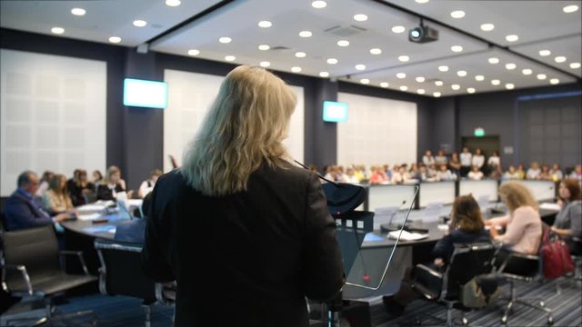 Modern adult woman make hand gesture during a conference at business forum. Woman explains to audience important information at symposium. Rear view of woman and microphone in audience at round table.