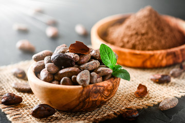 Cocoa beans with mint on jute side view. Cacao powder in wooden bowl blure in background. Panorama or banner concept.