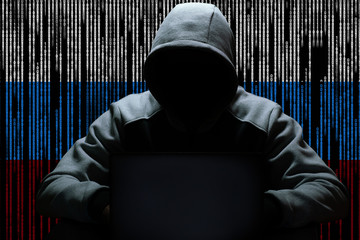 A Russian hacker is sitting in the dark in a hood in front of a computer, against a background of a...