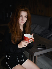 Cute redhead woman woman with adorable smile sitting in cozy city cafe enjoying free time coffee break holding big red mug in hands dressed black blouse and shorts.